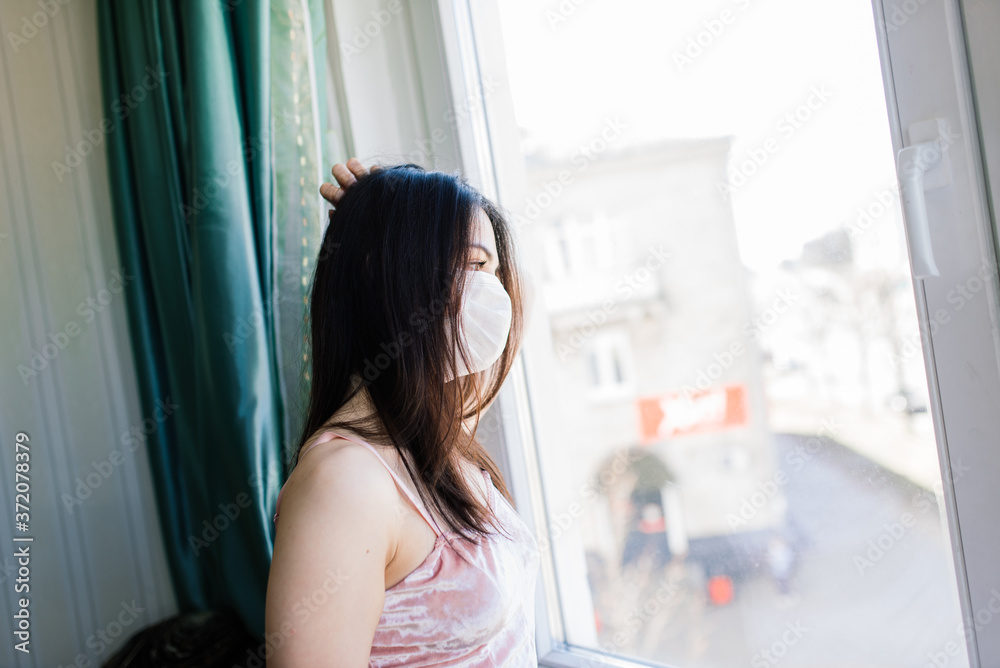 Sad lonely girl isolated at home in protective sterile medical mask on her face looking at window, bored woman because of Chinese pandemic coronavirus, virus covid-19.