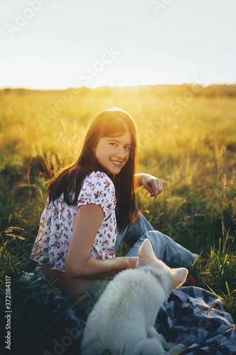 Happy woman sitting with cute white puppy in summer meadow in sunset warm light. Stylish girl relaxing with her adorable fluffy puppy on a picnic. Loyal friend. Summer travel with pet