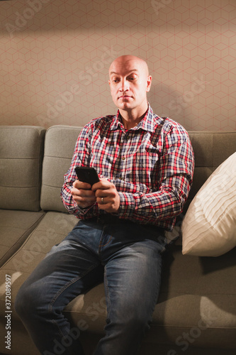Portrait of a happy adult bald man, dressed in a plaid shirt and jeans, sitting on the couch with a phone. © Евгений Гончаров