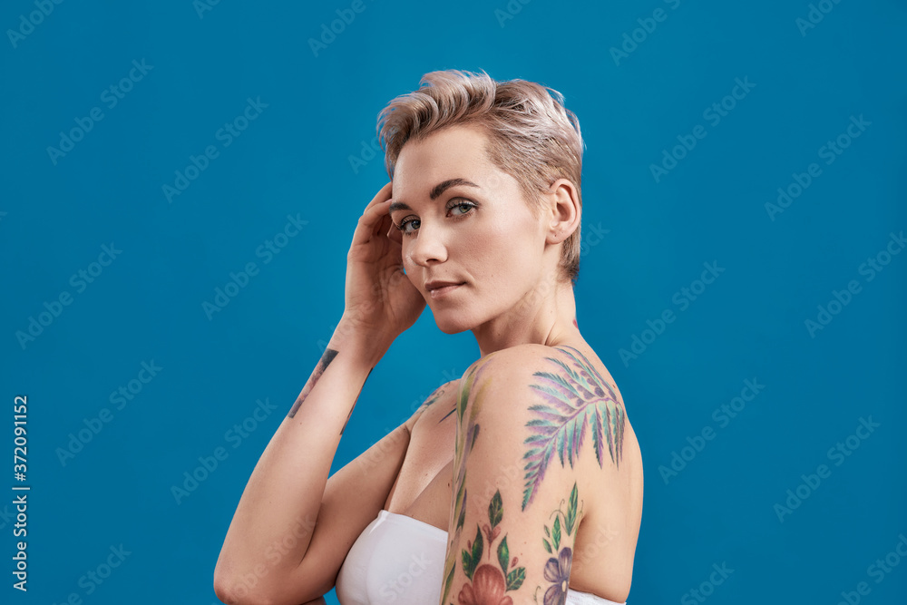 Beauty portrait of a young attractive half naked tattooed woman with perfect skin looking at camera, holding hand near her head isolated over blue background