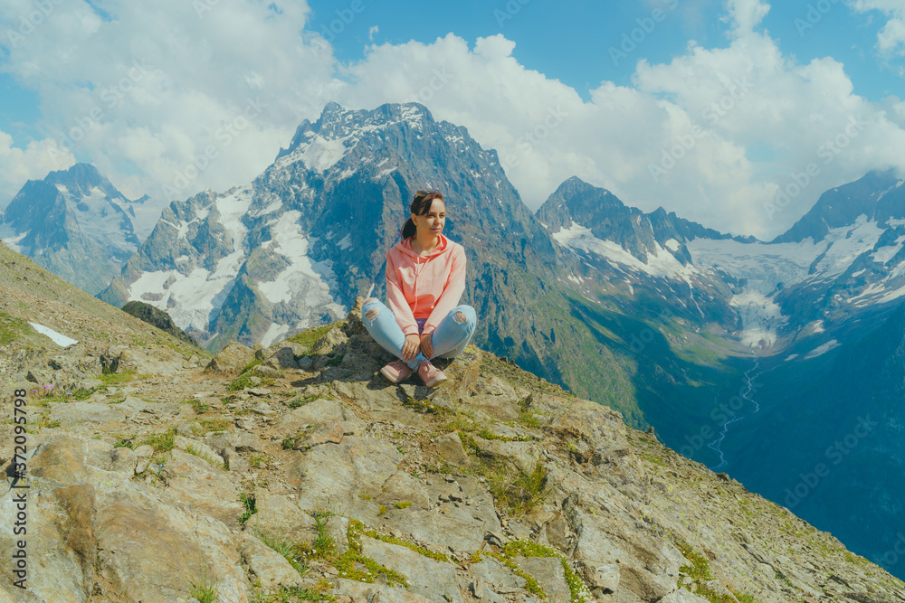 Young woman sitting on rock and looking at mountain landscape. Female traveler enjoying beautiful view in mountainous area.