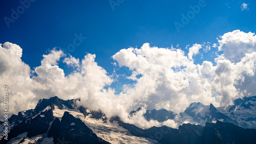 Snowy mountain against cloudy sky. From below white clouds floating on blue sky over mountain ridge covered with snow on sunny day in nature