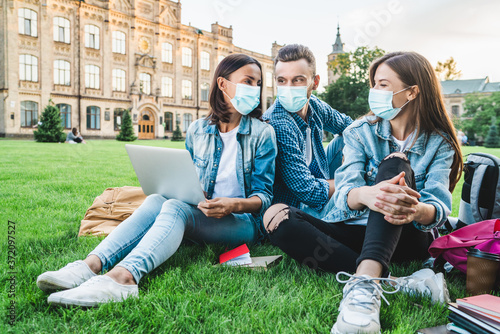 Group of young students studying outdoors during the quarantine preparing to finals while sitting on a grass lawn wearing medical masks for protection against virus