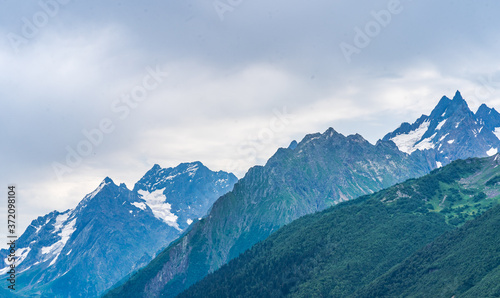 Beautiful mountain landscape in wooded area in summertime. Mighty mountains with snow and green array in cloudy weather.