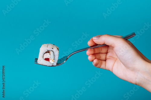 Hand holding fresh sushi roll with a fork, isolated on blue background. How to eat sushi without chopsticks