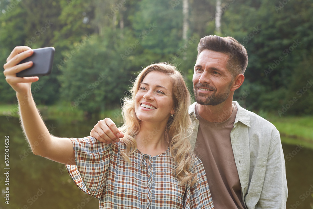 Waist up portrait of beautiful adult couple taking selfie while standing by lake in green countryside scenery, copy space