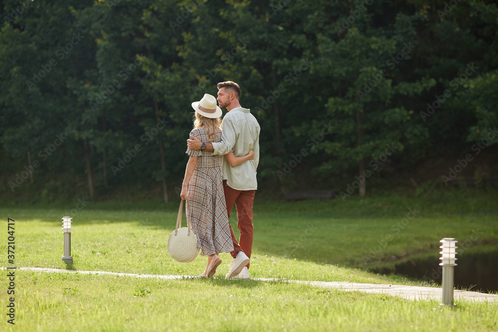 Back view full length portrait of romantic adult couple embracing while walking on path across green lawn in nature scenery, copy space