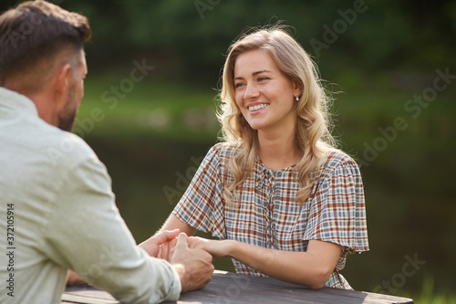 Portrait of beautiful couple holding hands and looking at each other with love while sitting at outdoor table by lake during romantic date