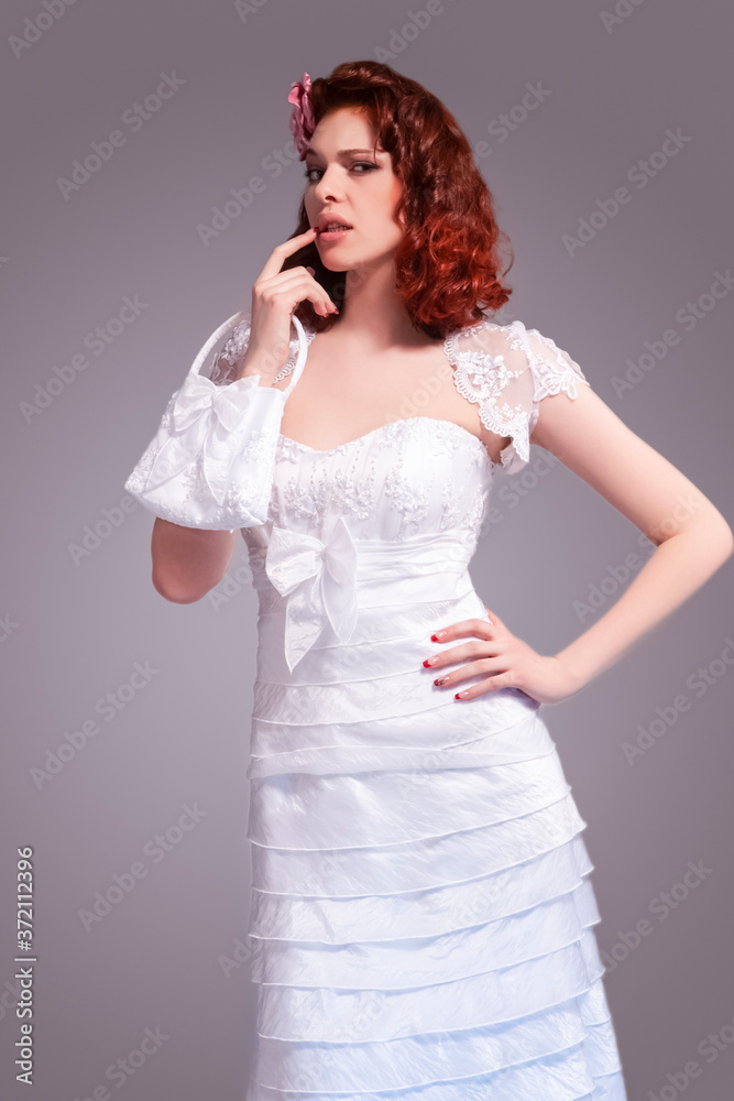 Portrait of Sad Redhaired Caucasian Female Posing in White Wedding Dress and Little Purse Against Gray Background. Portrait of Sad Redhaired Caucasian Female Posing in White Wedding Dress and Little Purse Against Gray Background.