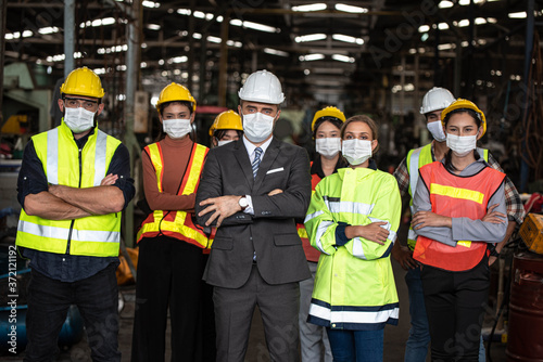 Group of industrial staff worker consist of Technicians, Engineers and factory Manager wearing helmet or hardhat with face mask to protect Covid 19 inflection in Industry plant/Standing post Teamwork