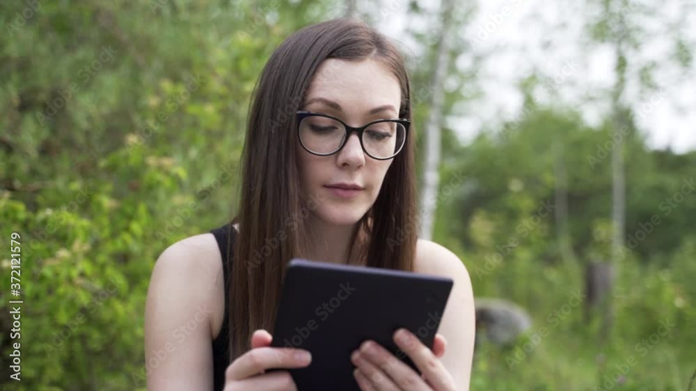 Young woman with glasses sitting in nature, reading on e-reader and smiling into camera, close up Young woman with glasses sitting in nature, reading on e-reader and smiling into camera, close up