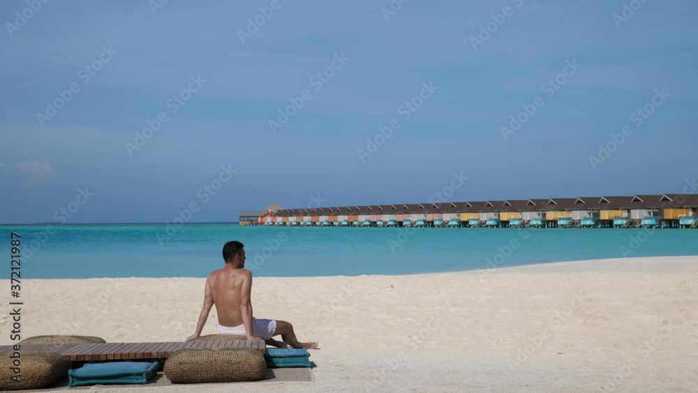 Caucasian male relaxes on beach alone with colourful villas stretching to the horizon Caucasian male relaxes on beach alone with colourful villas stretching to the horizon