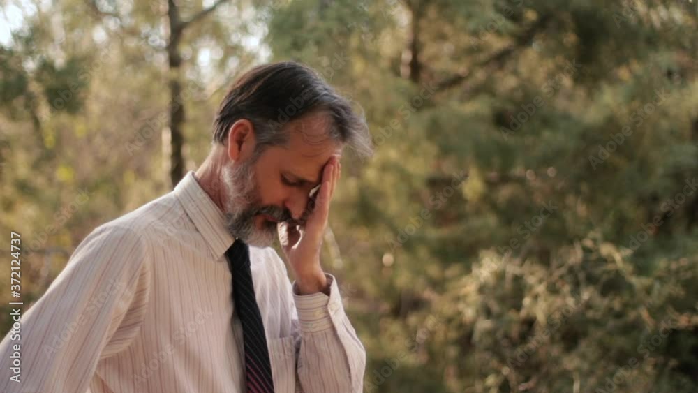 Tired, lonely, middle aged European bearded man yawns, touches face in outdoor park Tired, lonely, middle aged European bearded man yawns, touches face in outdoor park