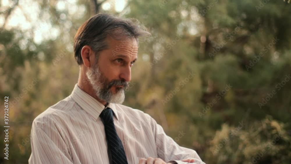 Suspicious old man in formal clothes checks watch and looks around stealthily in park Suspicious old man in formal clothes checks watch and looks around stealthily in park