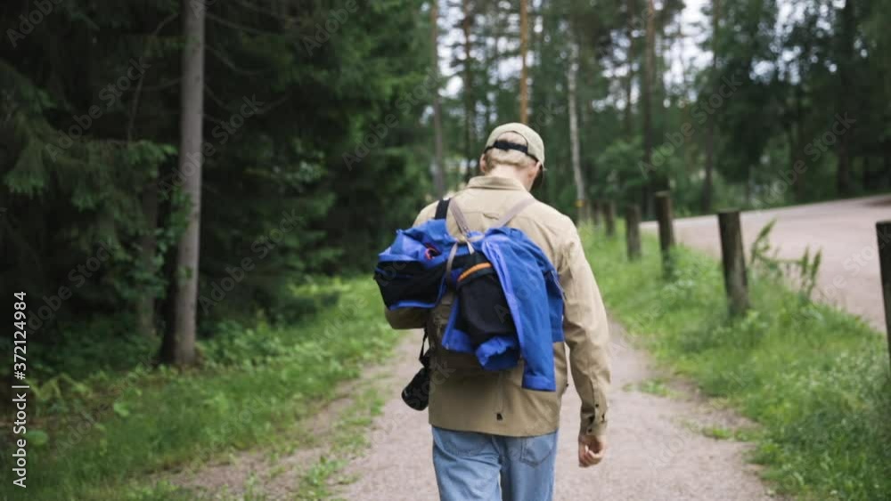 Young man with backpack and camera walks on gravel path by forest Young man with backpack and camera walks on gravel path by forest
