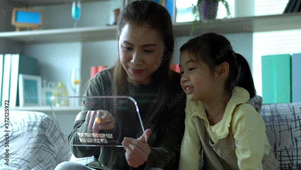 A futuristic virtual simulation of an Asian beautiful mother and a young daughter enjoy playing with an advance tablet mock up and answering an incoming video call at home A futuristic virtual simulation of an Asian beautiful mother and a young daughter enjoy playing with an advance tablet mock up and answering an incoming video call at home