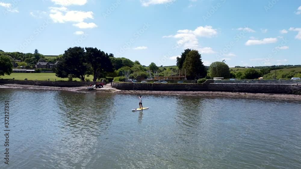 Drone pull out shot of male paddle boarder slowing paddling through the water in the summer sunshine. Drone pull out shot of male paddle boarder slowing paddling through the water in the summer sunshine.