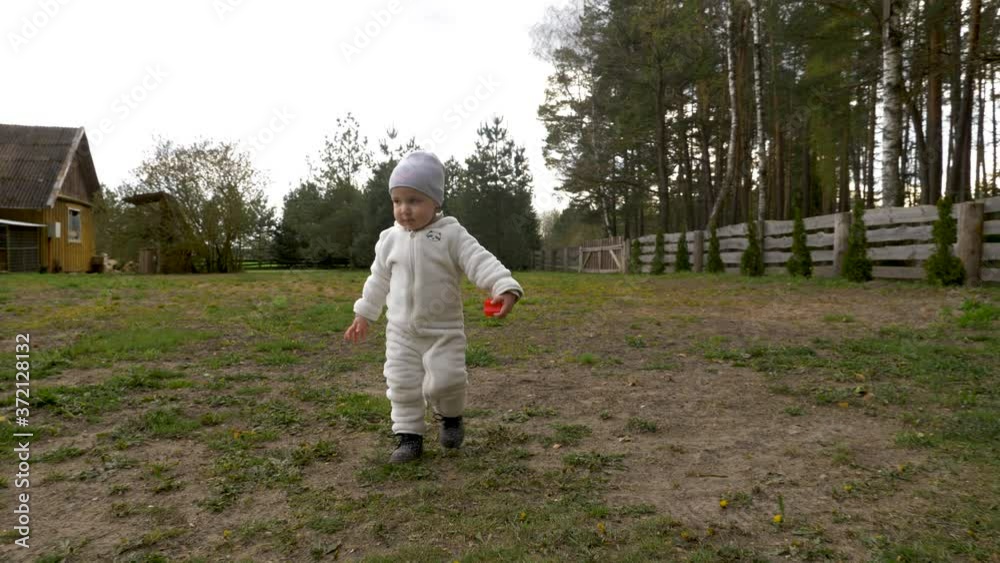 Toddler girl wearing warm clothes, walks in the yard with her toys. Little smiling girl walks straight to the camera. Toddler girl wearing warm clothes, walks in the yard with her toys. Little smiling girl walks straight to the camera.