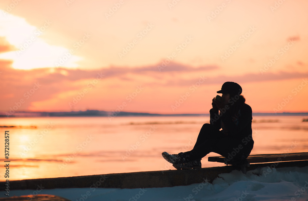 A girl sits on a bench against the background of the sunset sky and the river and drinks coffee from a cup.