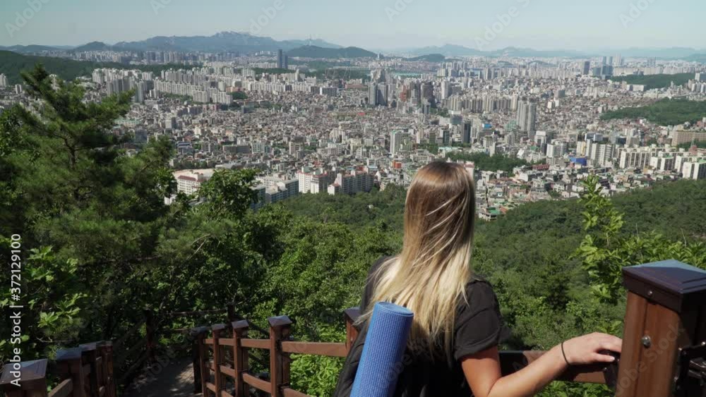 Blonde Girl Leaning On The Fence And Looking Towards The Beautiful City Of Seocho-gu District With High Rise Buildings From The Gwanaksan Mountain Trail In Seoul, South Korea. - wide shot Blonde Girl Leaning On The Fence And Looking Towards The Beautiful City Of Seocho-gu District With High Rise Buildings From The Gwanaksan Mountain Trail In Seoul, South Korea. - wide shot