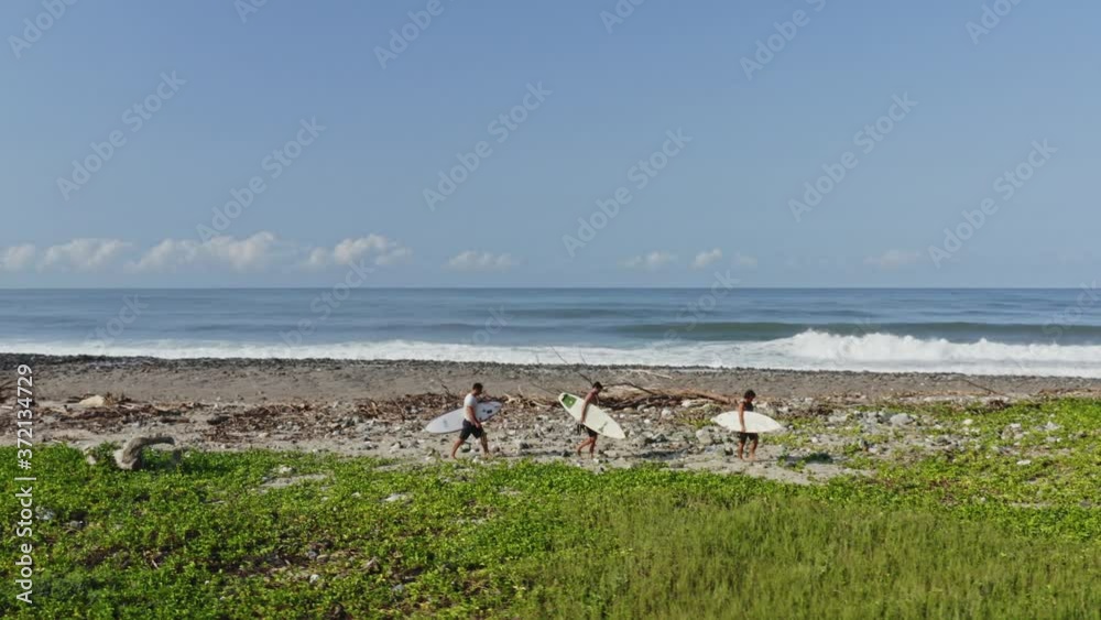 Aerial: surfers walking on island beach with surfboards, large waves backdrop Aerial: surfers walking on island beach with surfboards, large waves backdrop