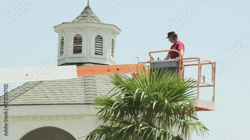 Young Caucasian skillful man power washing outdoor gazebo roof using orange industrial lift equipment to spray and clean roof, static low angle photo