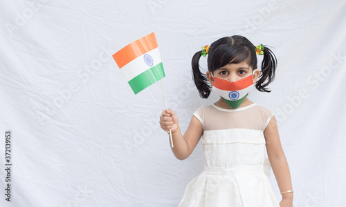 Independence Day (August 15) - Indian girl in white dress with braided hair, wearing tri color face mask of Indian flag and holding Indian national flag