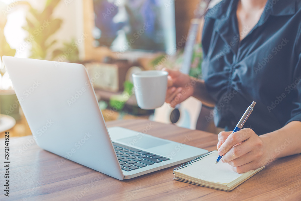 Woman is writing on a notebook with a pen and using a laptop to work in the office.