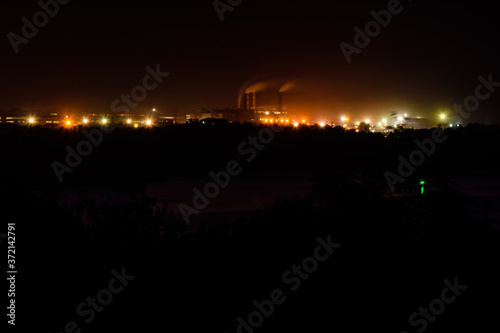 View on city lights at night. Smoke from chimneys of the factory rises to sky. Environmental pollution concept