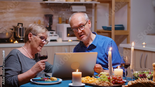 Aged, retired, couple shopping online during romanitic dinner using laptop. Old people sitting at the table, browsing, using the technoloty, internet, celebrating their anniversary in the dining room. photo