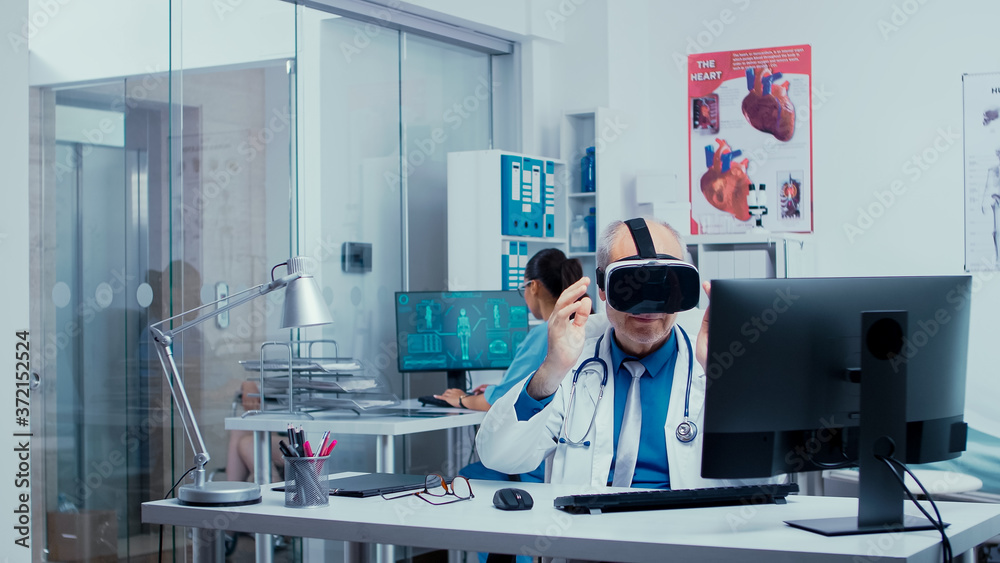 Senior physician using VR glasses in modern private clinic to study diseases in virtual space and modern technology. In the background modern clinic with glass walls and patients with doctors in Senior physician using VR glasses in modern private clinic to study diseases in virtual space and modern technology. In the background modern clinic with glass walls and patients with doctors in