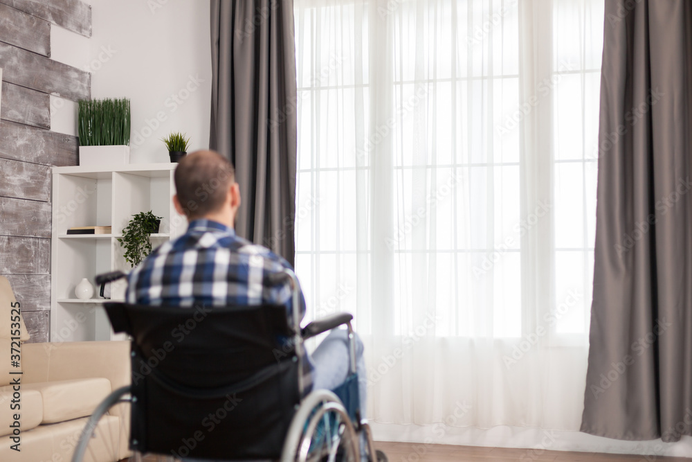 Lonely man in wheelchair with walking disability in living room. Serious sad caucasian man wearing casual clothes and look at large panoramic view in bright modern living room, hospital or clinic. Lonely man in wheelchair with walking disability in living room. Serious sad caucasian man wearing casual clothes and look at large panoramic view in bright modern living room, hospital or clinic.