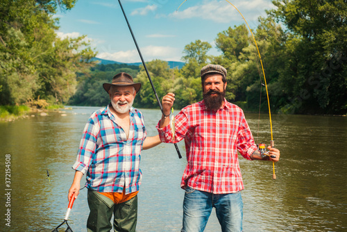 Activity and hobby. Set up rod with hook line sinker. Two male friends dressed in shirts fishing together with net and rod during the morning light on lake. Portrait of cheerful senior man fishing.