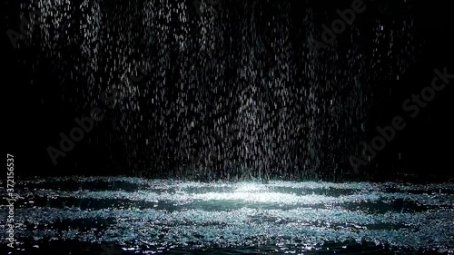 Spray spatters rain fall against on the water surface which glow. Black background. White neon lighting in dark studio. Slow motion. photo