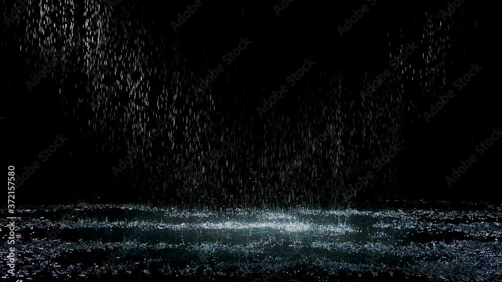 Spray spatters rain fall against on the water surface which glow. Black background. White neon lighting in dark studio. Slow motion.