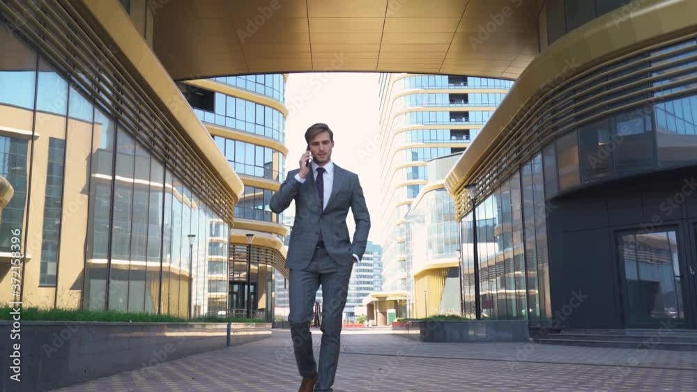 Young businessman in a suit walking next to the glass skyscraper and talking on a mobile phone, top manager near the modern office. Young businessman in a suit walking next to the glass skyscraper and talking on a mobile phone, top manager near the modern office.
