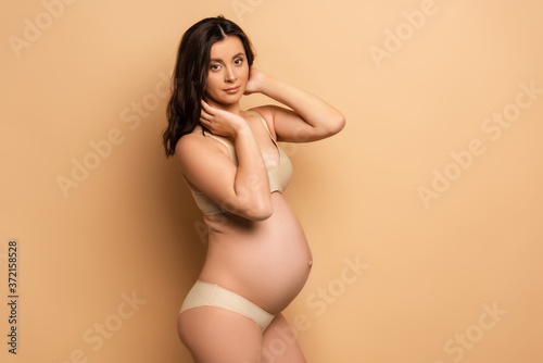  pregnant brunette woman in underwear touching hair while looking at camera on beige
