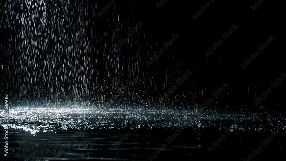 From the ceiling spatters rain fall against on the water surface which glow. Black background. White neon lighting in dark studio. Slow motion. Close up.