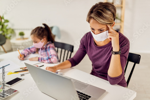 Working mother using laptop while her daughter is doing homework during coronavirus epidemic.