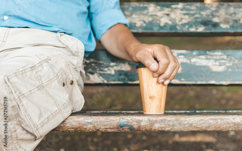 Cropped image of senior man sitting on park bench and holding reusable or easily recyclable bamboo cup for take away coffee. Tranquillity, relax and sustainable lifestyle concept.