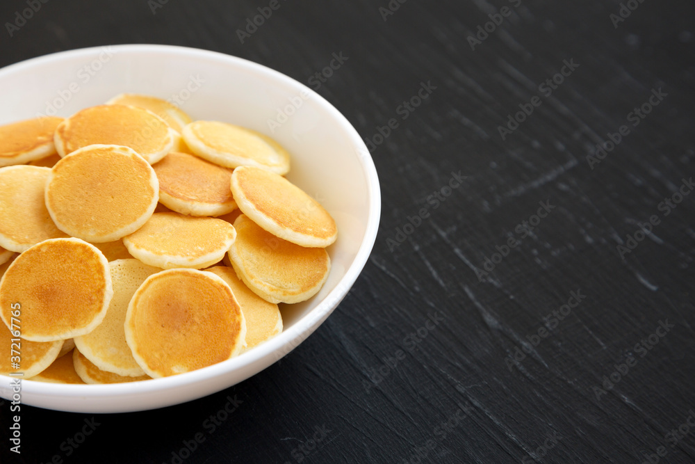 Homemade Mini Pancakes Cereal in a white bowl on a black background, side view. Copy space. Homemade Mini Pancakes Cereal in a white bowl on a black background, side view. Copy space.