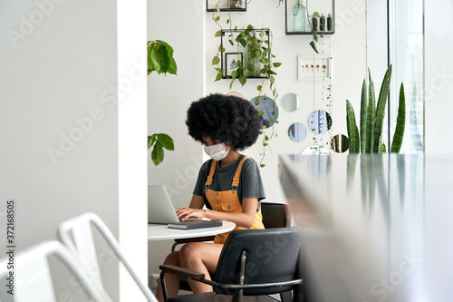 Young African American teen girl student with Afro hair wearing face mask using laptop sitting at cozy cafe table alone indoor. Social distancing and work, study on computer with covid 19 protection.