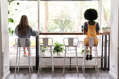 Two diverse young women sitting apart at cafe table, African and Caucasian girls working or studying, dining in cafeteria avoiding communication, social distancing safety indoors concept, back view. photo