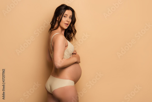 pregnant brunette woman in underwear looking at camera while touching belly on beige background