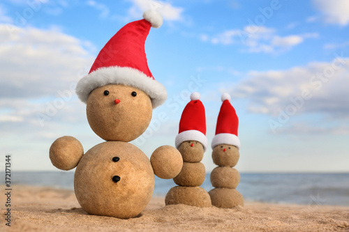 Snowmen made of sand with Santa hats on beach near sea. Christmas vacation