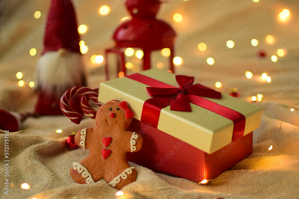 New Year's Christmas decoration. Christmas tree toy in the form of a cookie near a gift box with a red bow on the background of a soft beige plaid. In the background a handmade gnome and a red lantern New Year's Christmas decoration. Christmas tree toy in the form of a cookie near a gift box with a red bow on the background of a soft beige plaid. In the background a handmade gnome and a red lantern