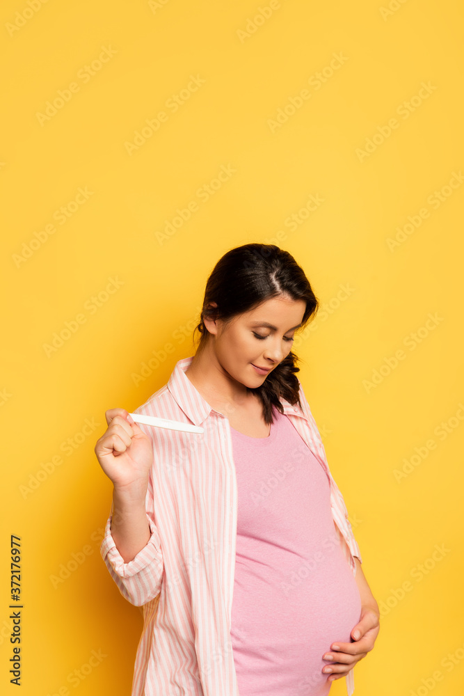 pregnant woman holding pregnancy test while touching tummy on yellow pregnant woman holding pregnancy test while touching tummy on yellow