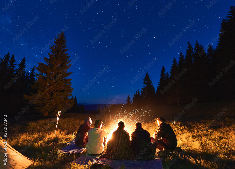 Summer camping under stars. Back view of group of five people, men and woman sitting near bright bonfire, tourist tent under dark night sky with sparkling stars. Concept of tourism, evening camping. - obrazy, fototapety, plakaty 