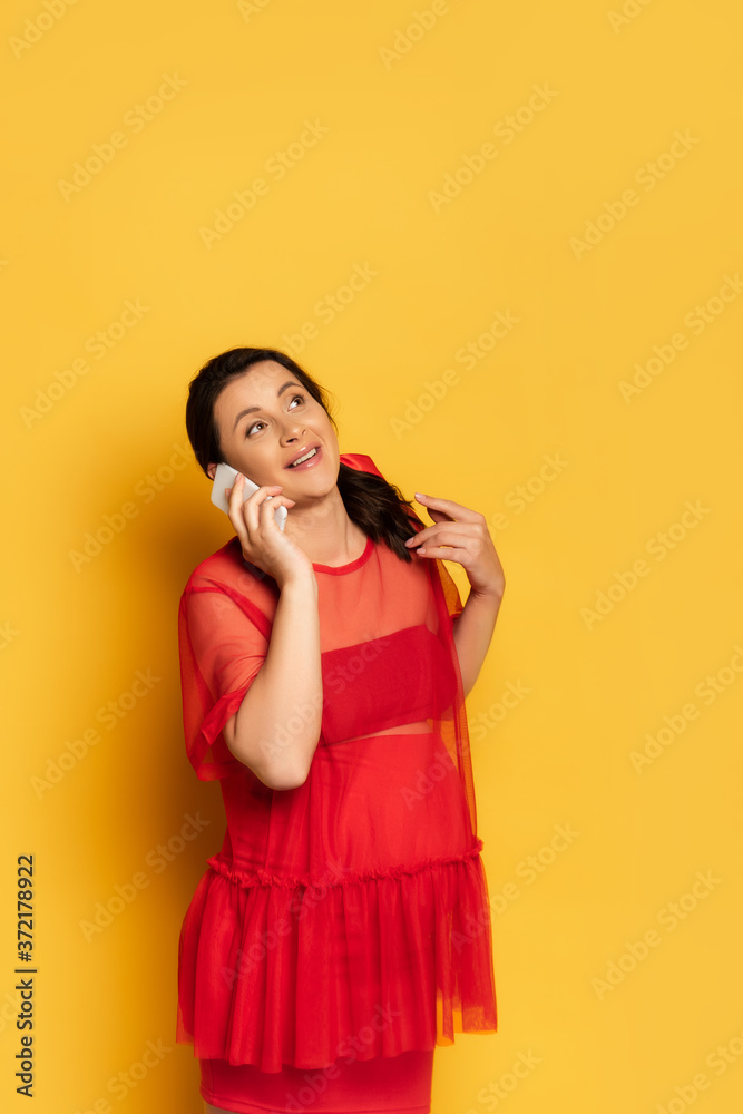 Fototapeta premium pregnant woman in red tunic looking up while talking on smartphone on yellow