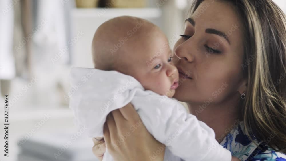 delighted young mother with long loose hair holds and kisses little baby in white bodysuit and pulls faces playing with kid at home close view delighted young mother with long loose hair holds and kisses little baby in white bodysuit and pulls faces playing with kid at home close view